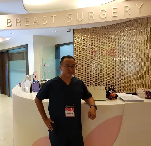 BS An mặc trang phục phẫu thuật, đeo thẻ tham dự, đứng tại quầy tiếp đón của THE Plastic Surgery Clinic, Seoul