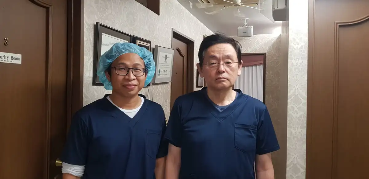 BS An đội mũ phẫu thuật, chụp ảnh cùng một bác sĩ đồng nghiệp, cả hai mặc trang phục phẫu thuật, tại Nhật Bản